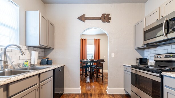 A kitchen with a wooden floor, stainless steel appliances, and a dining area with orange curtains. - The Cozy Chickasha | Fiber | Extra Parking | W/D (Chickasha)