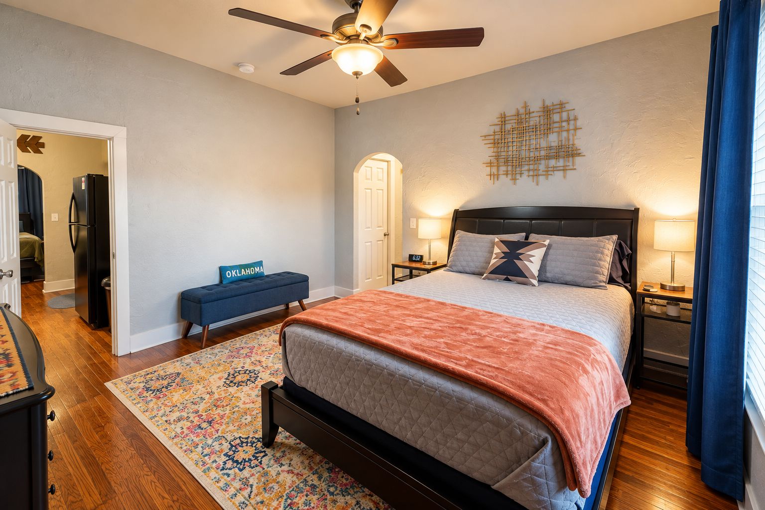 Quarto com cama, ventilador de teto, mesa de cabeceira com luminária, um banco e decoração na parede.
