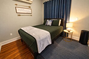Un lit bien fait, avec une couvre-lit verte et un plaid blanc, une table de chevet avec une lampe et une poubelle.