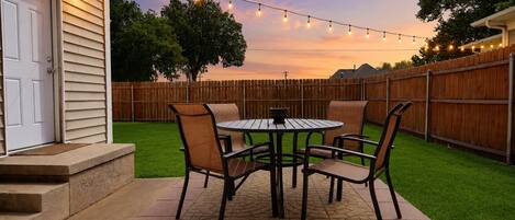 A patio with a round table and chairs, a wooden fence, and string lights.