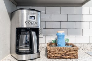 A kitchen with a stainless steel coffee maker, a basket with jars, and a water bottle on the counter.