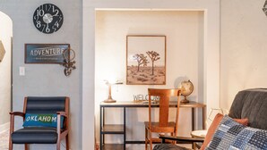 A living room with a wooden desk, a chair with a pillow labeled "OKLAHOMA," a clock, a framed picture of a desert landscape, and a couch with patterned cushions. - The Cozy Chickasha | Fiber | Extra Parking | W/D (Chickasha)