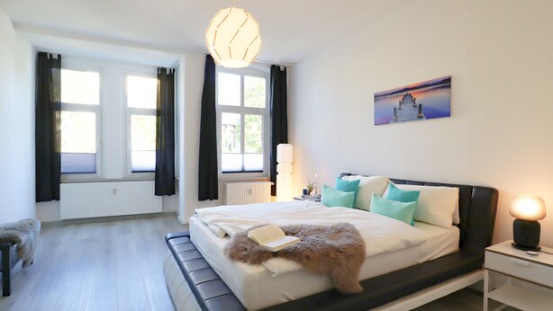 A modern bedroom with a large bed, a bedside table with a lamp, a radiator, and a window with curtains. - *NEW* Central (5min to city center) *Netflix & Amazon Prime* Kleiststrasse 4 MD (Sachsen-Anhalt - Magdeburg)