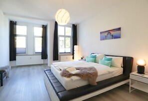 Ein modernes Schlafzimmer mit einem großen Bett, einem Nachttisch mit Lampe, einem Heizkörper und einem Fenster mit Vorhängen.