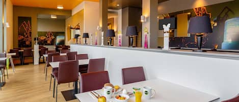 Un comedor moderno con varias mesas y sillas, una barra con una máquina de café y diversas opciones de desayuno sobre las mesas.
