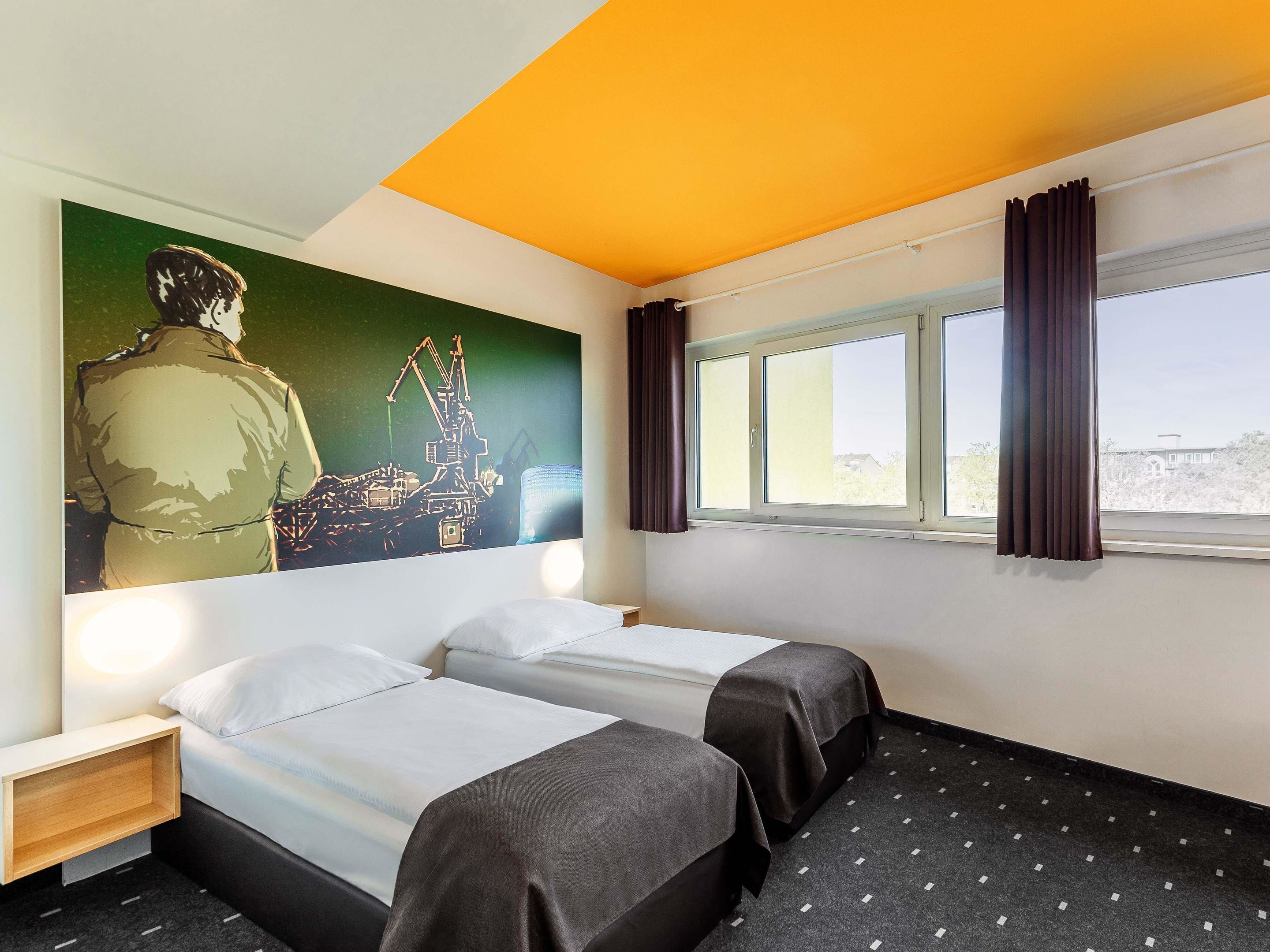 A hotel room with two beds, a large mural on the wall, a window with curtains, and a view of a building outside.