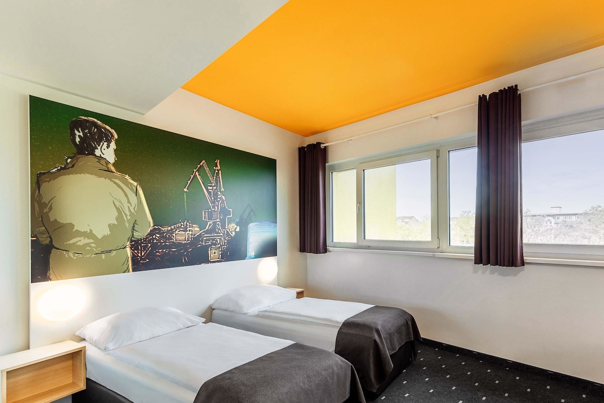 Une chambre d’hôtel avec deux lits, une grande fresque murale, une fenêtre avec des rideaux et une vue sur un bâtiment à l’extérieur.