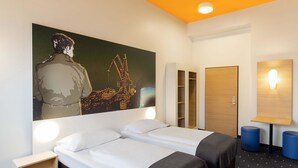 Une chambre d’hôtel comprenant un lit, deux tables de chevet, une armoire, un tableau au mur et un bureau avec une lampe.