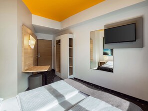Une chambre d’hôtel moderne équipée d’un lit, d’un bureau, d’une armoire et d’un téléviseur fixé au mur.