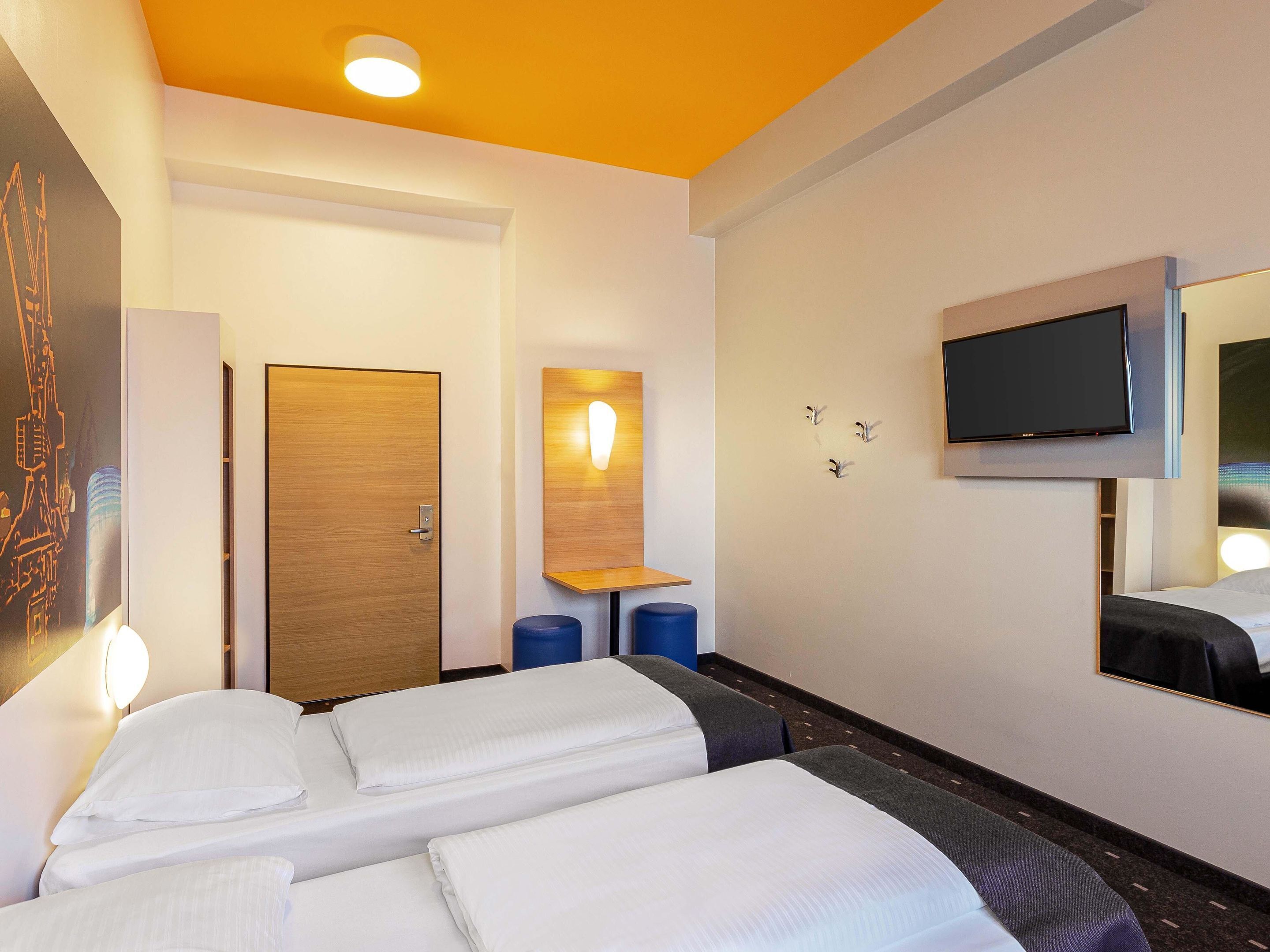 Ein Hotelzimmer mit zwei Betten, einer Holztür, einem an der Wand befestigten Fernseher und einem kleinen Regal.