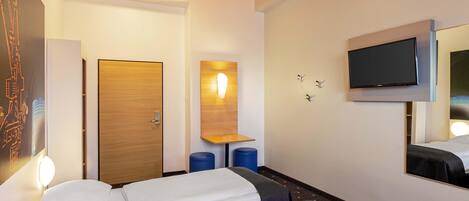 Habitación de hotel con dos camas, una puerta de madera, un televisor montado en la pared y un pequeño estante.