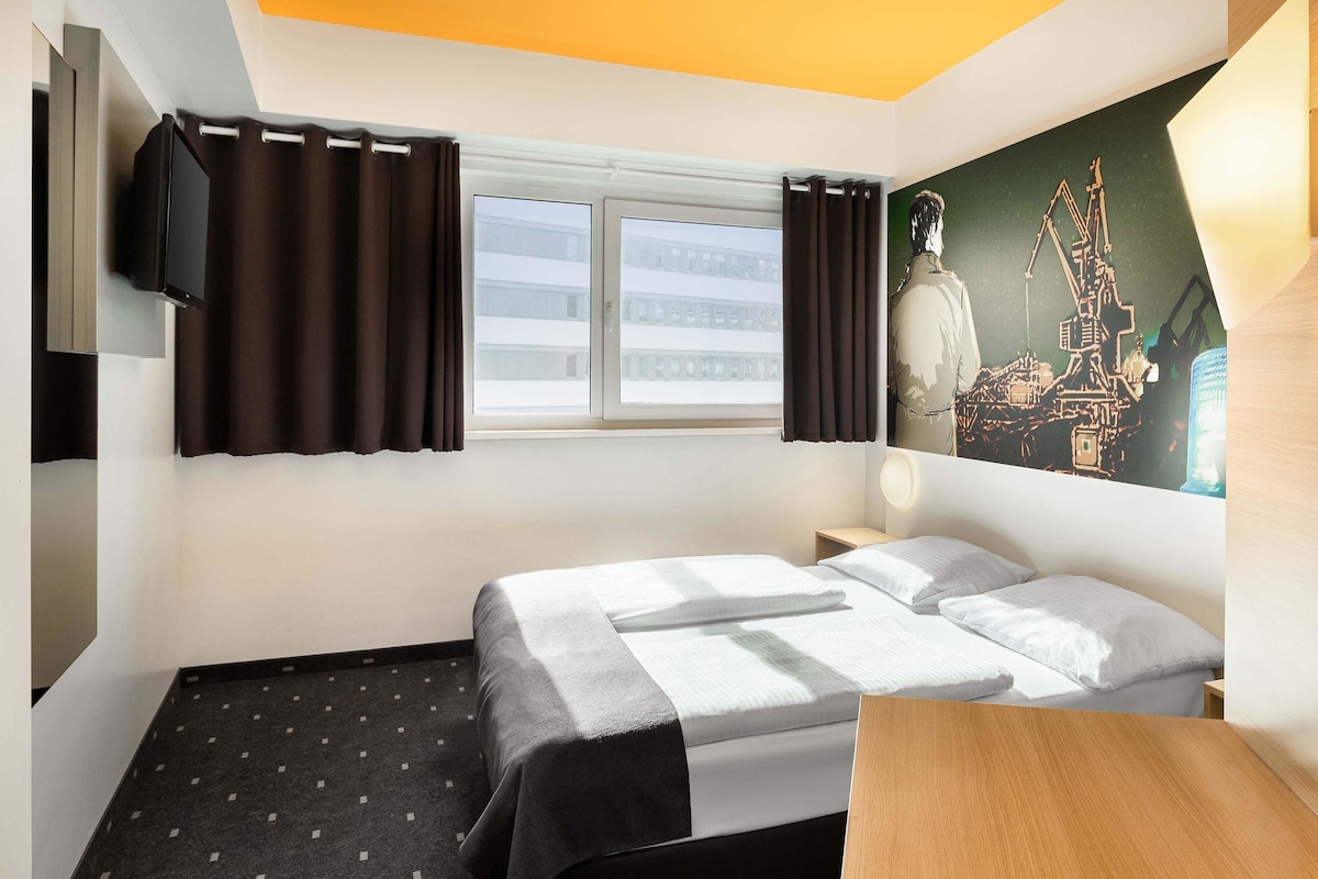 Une chambre d’hôtel comprenant un lit, un bureau, une fenêtre avec des rideaux, un téléviseur fixé au mur et un tableau représentant un paysage urbain.