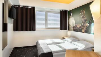 Ein Hotelzimmer mit Bett, Schreibtisch, einem Fenster mit Vorhängen, einem an der Wand befestigten Fernseher und einem Stadtbild.