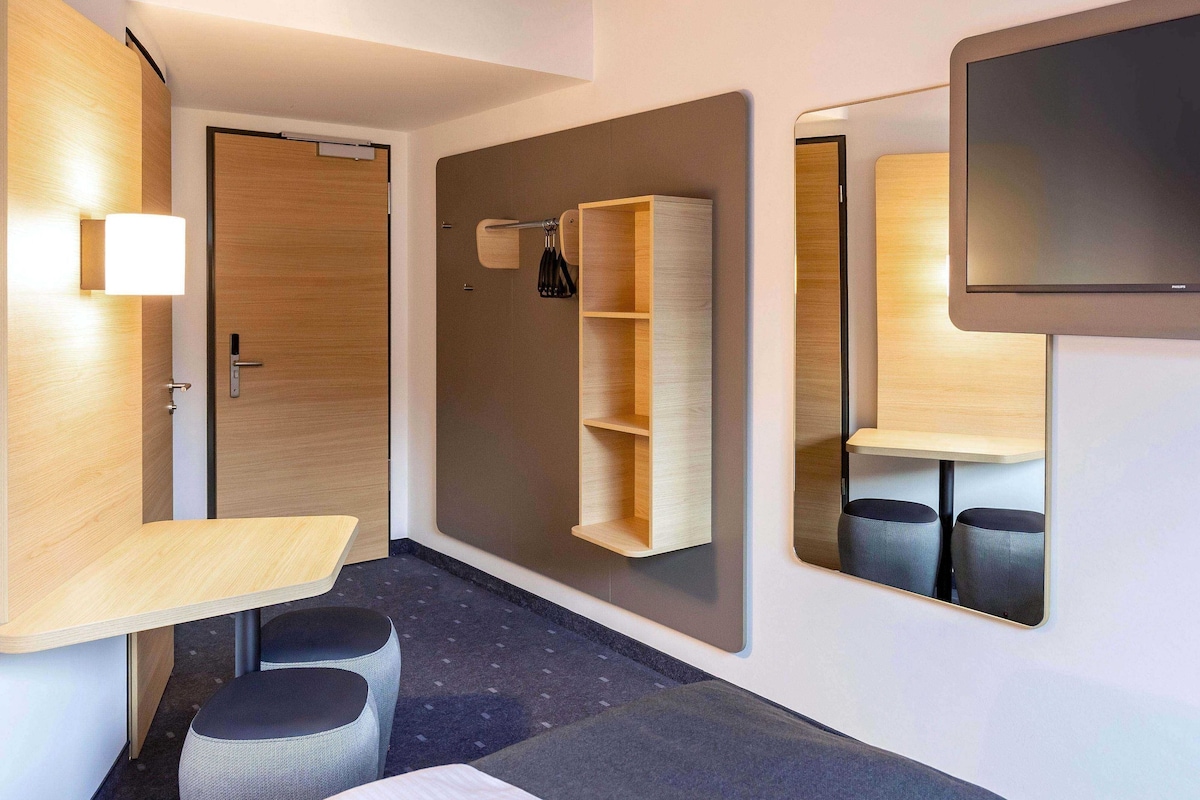 Une chambre d’hôtel moderne avec une porte en bois, un petit bureau avec une chaise, un miroir et une étagère fixée au mur.