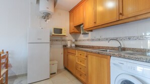 A compact kitchen with wooden cabinets, a white refrigerator, a microwave, a washing machine, and a tiled backsplash. - 040 - Gran Playa 001 - Santa Pola (Santa Pola)