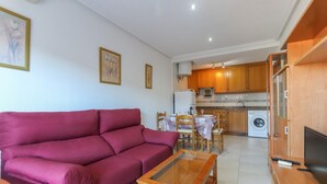 A living room with a red sofa, a wooden coffee table, and a TV stand. The kitchen area is visible in the background with wooden cabinets and a washing machine. - 040 - Gran Playa 001 - Santa Pola (Santa Pola)