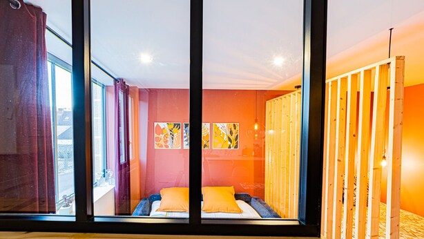 Un dormitorio moderno con un ventanal grande, una cama con almohadones naranjas y un separador de madera.