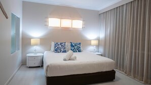 A neatly arranged bedroom with a bed, two bedside tables, a wall-mounted light fixture, and a window with curtains. - Poinciana Lodge 103 (Hamilton Island)