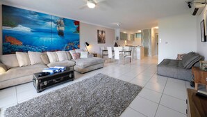 A spacious living room with a large ocean-themed painting, a dining area with white chairs, and a kitchen in the background. - Poinciana Lodge 103 (Hamilton Island)
