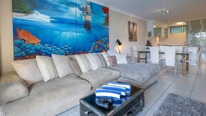 A living room with a large ocean-themed painting, a sofa, a coffee table with items, and a view into the kitchen. - Poinciana Lodge 103 (Hamilton Island)