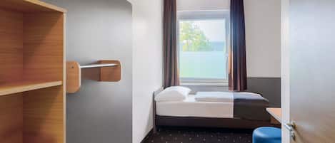 Una habitación de hotel pequeña con una cama, un estante de madera, una ventana con cortinas y un reposapiés azul.