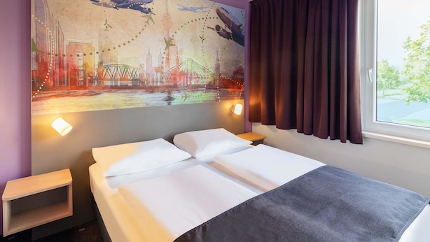 A hotel room with a bed, a nightstand, a wall-mounted lamp, and a window with curtains.