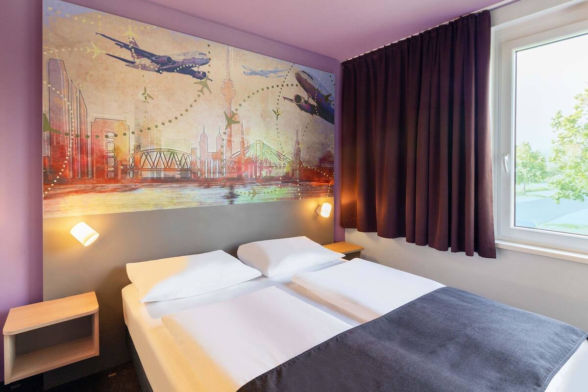 Una habitación de hotel con una cama, una mesita de noche, una lámpara de pared y una ventana con cortinas.