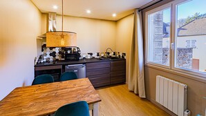 Cocina compacta con una mesa y sillas de madera, un lámpara colgante y vista a un edificio de piedra por una ventana.