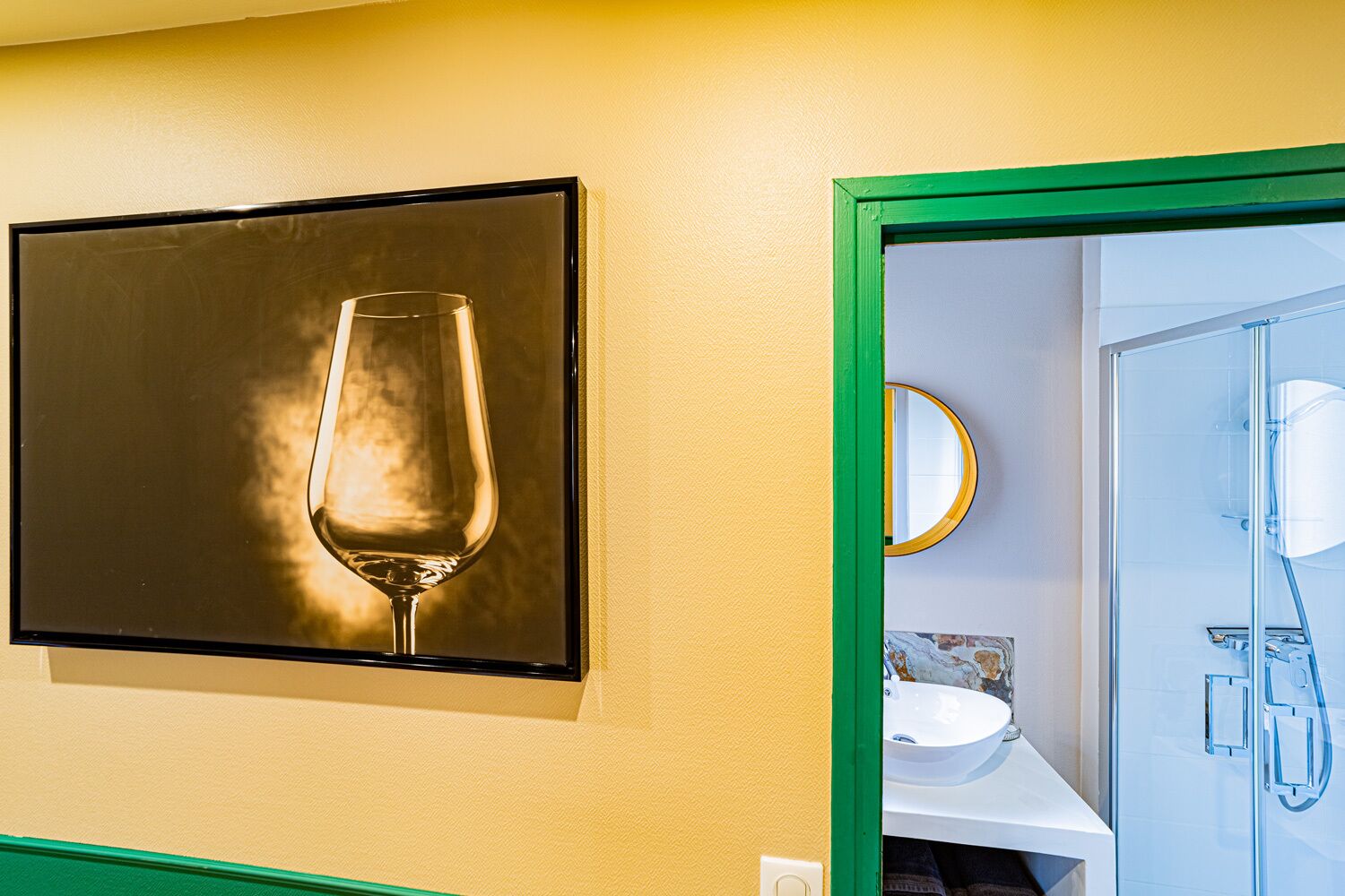 A framed artwork of a wine glass on a wall, a bathroom with a sink and mirror, and a shower area.