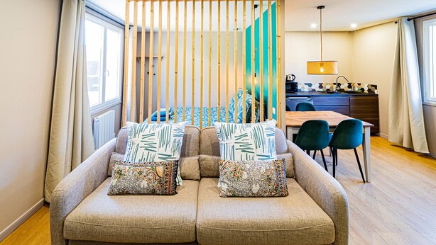 Una sala de estar moderna con un conjunto de sofá, cojines estampados, una zona de comedor con mesa y sillas, y una cocina al fondo.