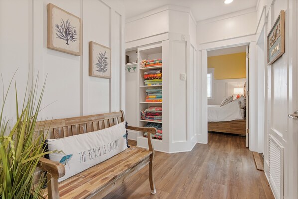 A wooden bench with a pillow, a white wall with framed artwork, a closet with folded blankets, and a bedroom with a bed and window. - Oceanfront Home with Expansive Views (Bald Head Island)
