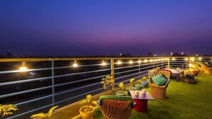 Terraza en la azotea con vistas al horizonte urbano al atardecer, con luces colgantes, plantas en macetas y mobiliario exterior.
