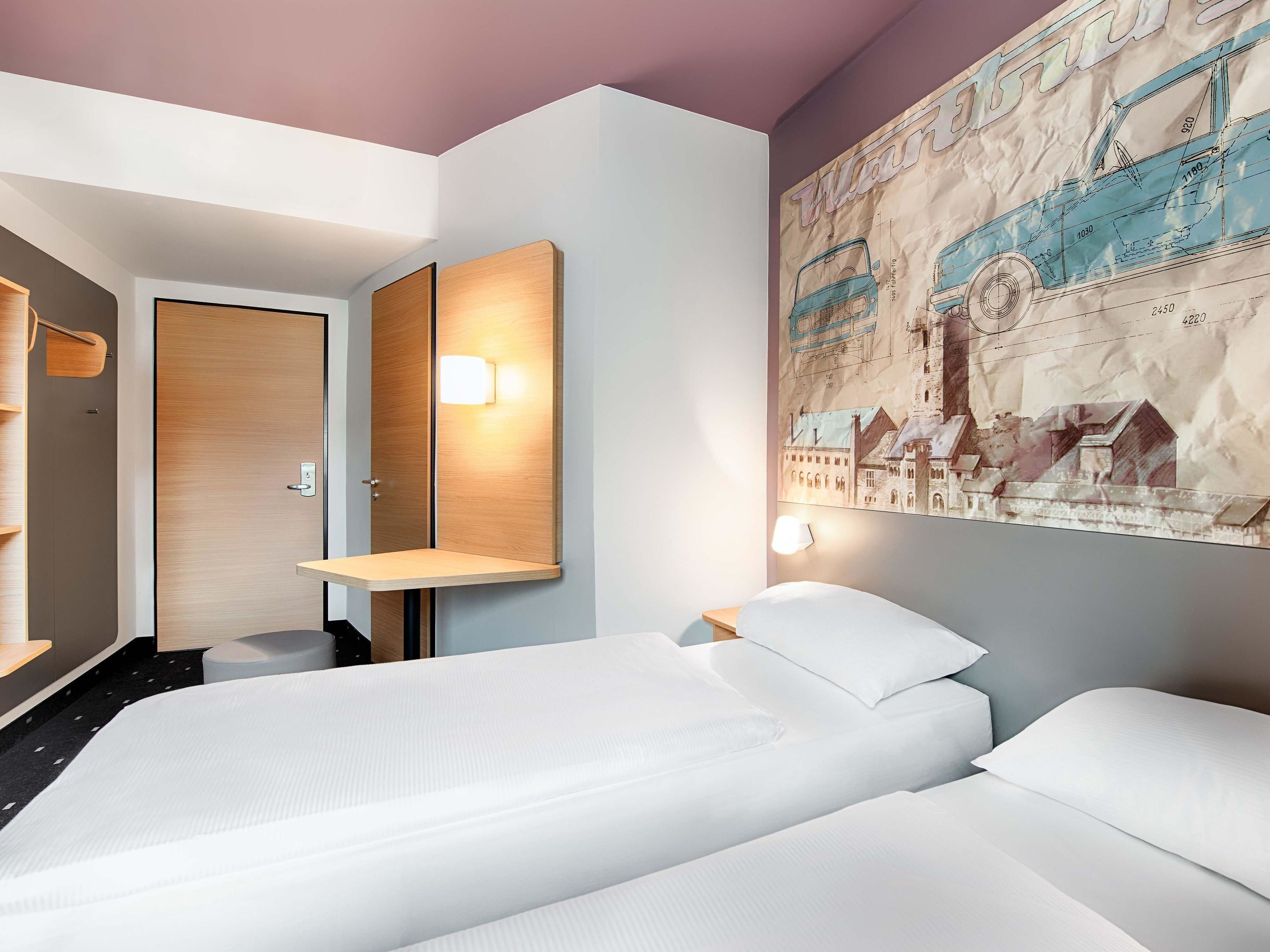 A hotel room with two beds, a wooden door, a wall-mounted mirror, and a large cityscape poster.