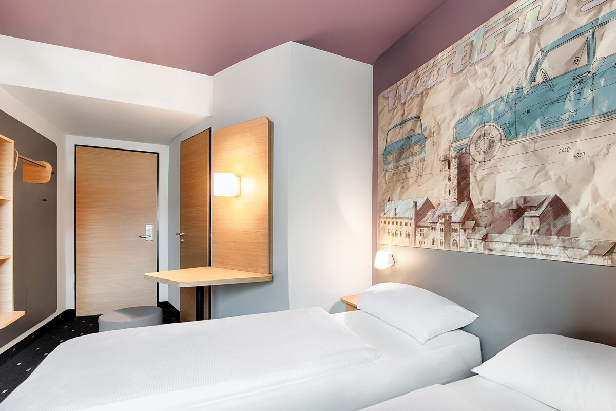 Ein Hotelzimmer mit zwei Betten, einer Holztür, einem wandmontierten Spiegel und einem großen Stadtplakats.