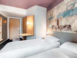 Une chambre d’hôtel avec deux lits, une porte en bois, un miroir fixé au mur et une grande affiche représentant un paysage urbain.