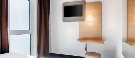 A hotel room with a bed, a television mounted on a wooden panel, a small table, and a trash bin.