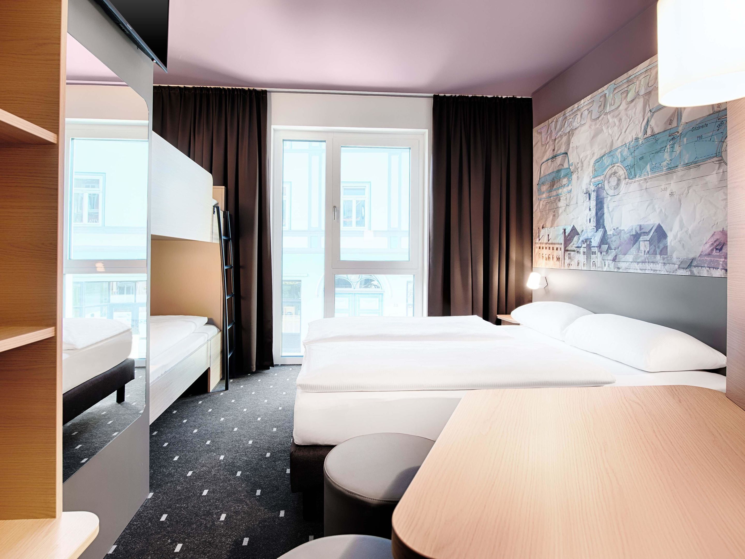 A hotel room with a bunk bed, a single bed, a desk, and a large window with curtains.