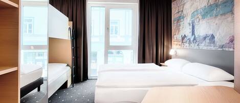 Une chambre d’hôtel avec un lit superposé, un lit simple, un bureau et une grande fenêtre avec des rideaux.
