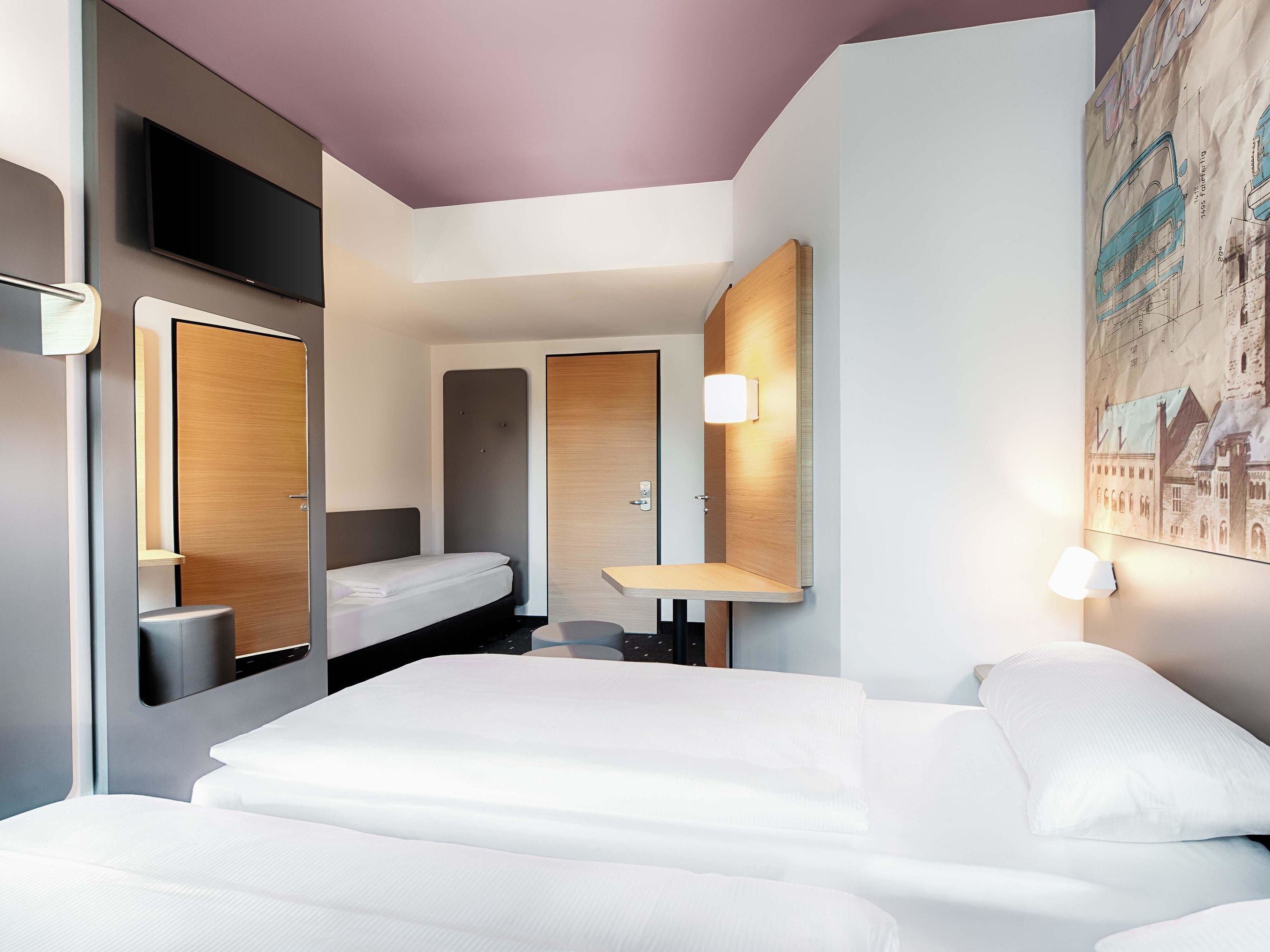 Une chambre d’hôtel moderne, équipée d’un lit, de tables de chevet, d’un miroir, d’un téléviseur fixé au mur et d’un tableau représentant un paysage urbain.