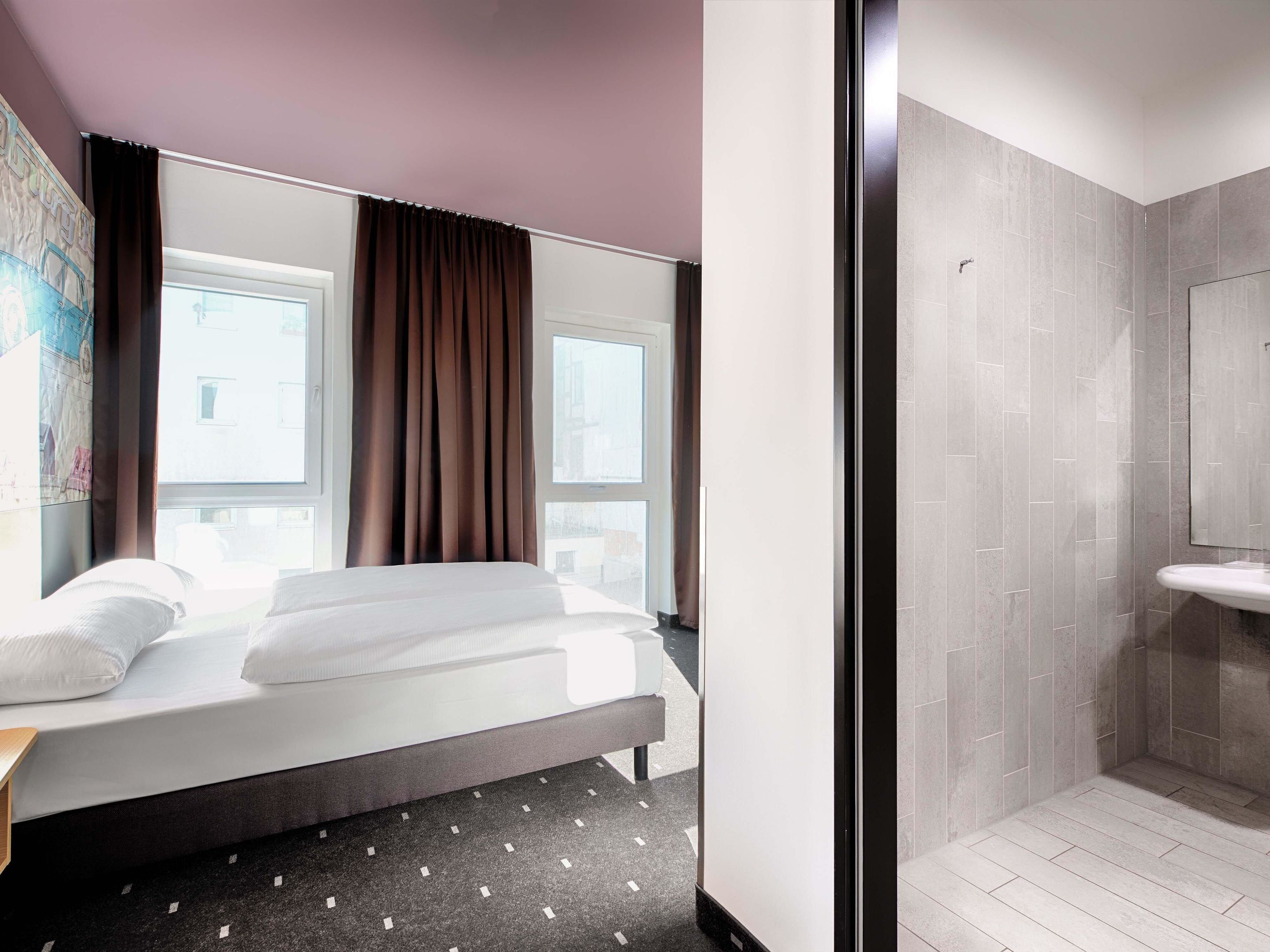 Ein Hotelzimmer mit Bett, Fenster mit Vorhängen sowie einem Badezimmer mit Waschbecken und gefliesten Wänden.