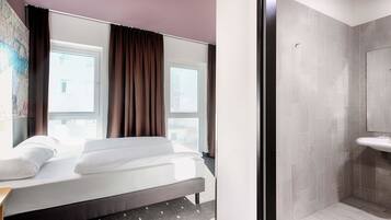 Ein Hotelzimmer mit Bett, Fenster mit Vorhängen sowie einem Badezimmer mit Waschbecken und gefliesten Wänden.