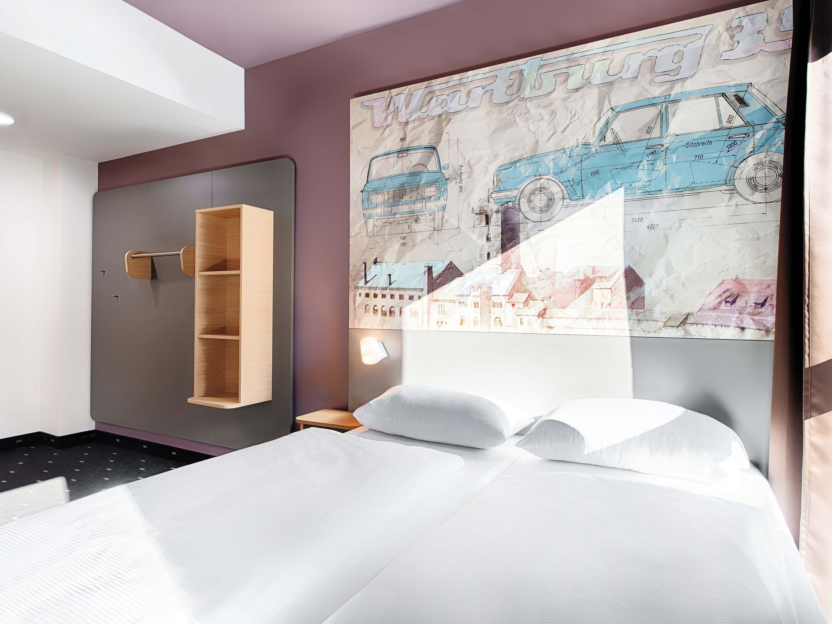 Ein Hotelzimmer mit Bett, einer Holzregaleinheit und einem großen Wandposter mit einem blauen Auto und Gebäuden.