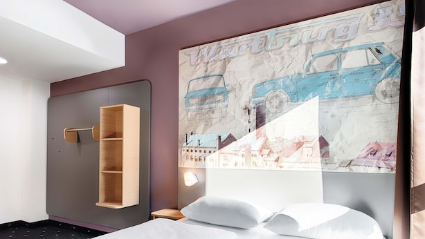 Habitación de hotel con cama, estantería de madera y un cartel grande en la pared con un coche azul y edificios.