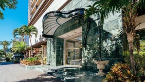 Entrada de hotel con fachada de mármol, un amplio arco decorativo y un jardín bien cuidado con plantas tropicales.
