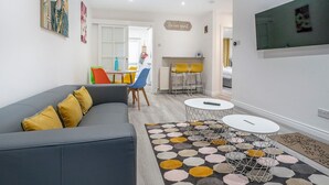 Una sala de estar moderna con un sofá gris, sillas coloridas, un comedor con mesa y sillas, y una cocina al fondo.