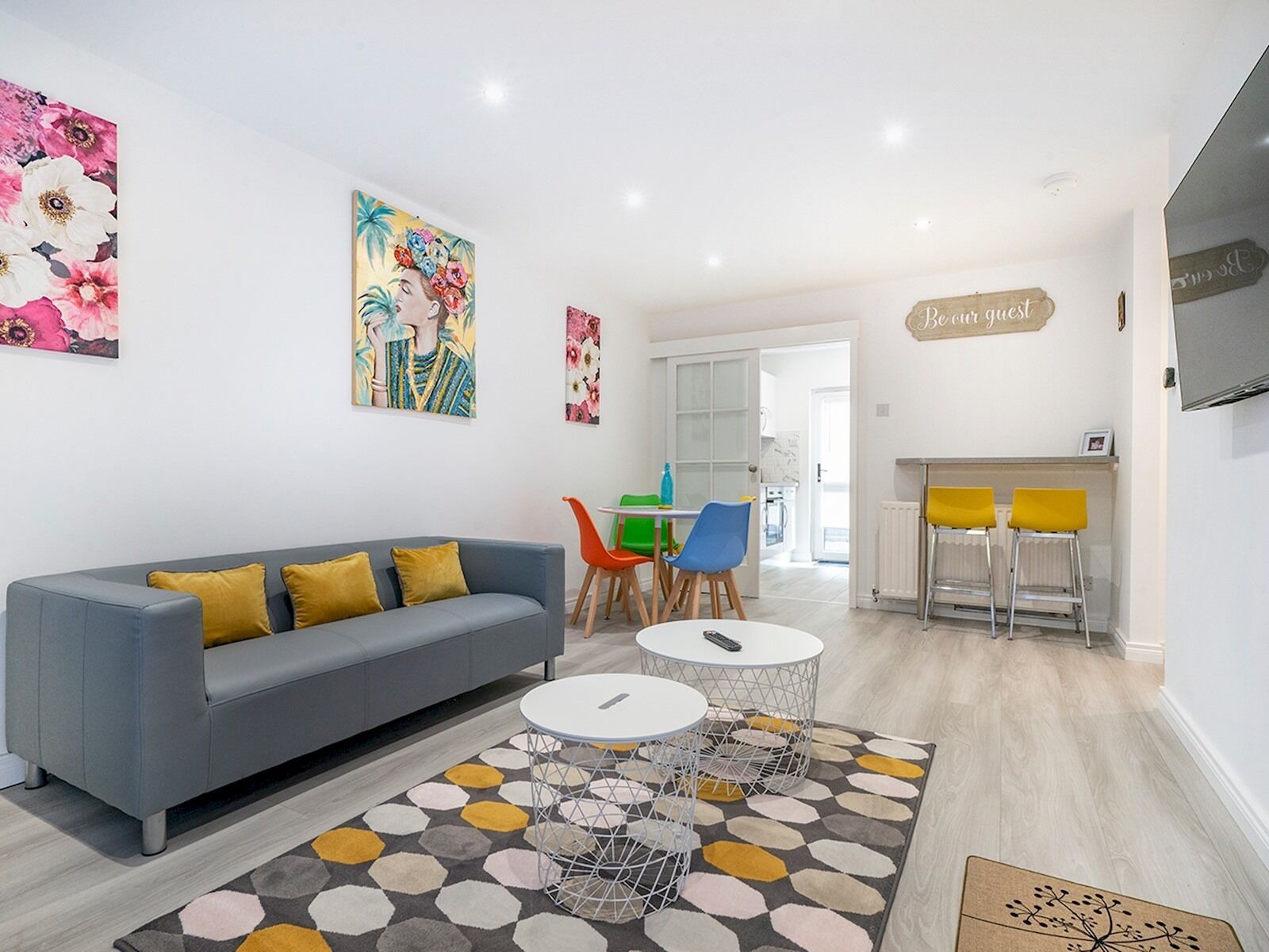 A modern living room with a grey sofa, colorful artwork, a dining area with yellow chairs, and a kitchen visible through an open door.