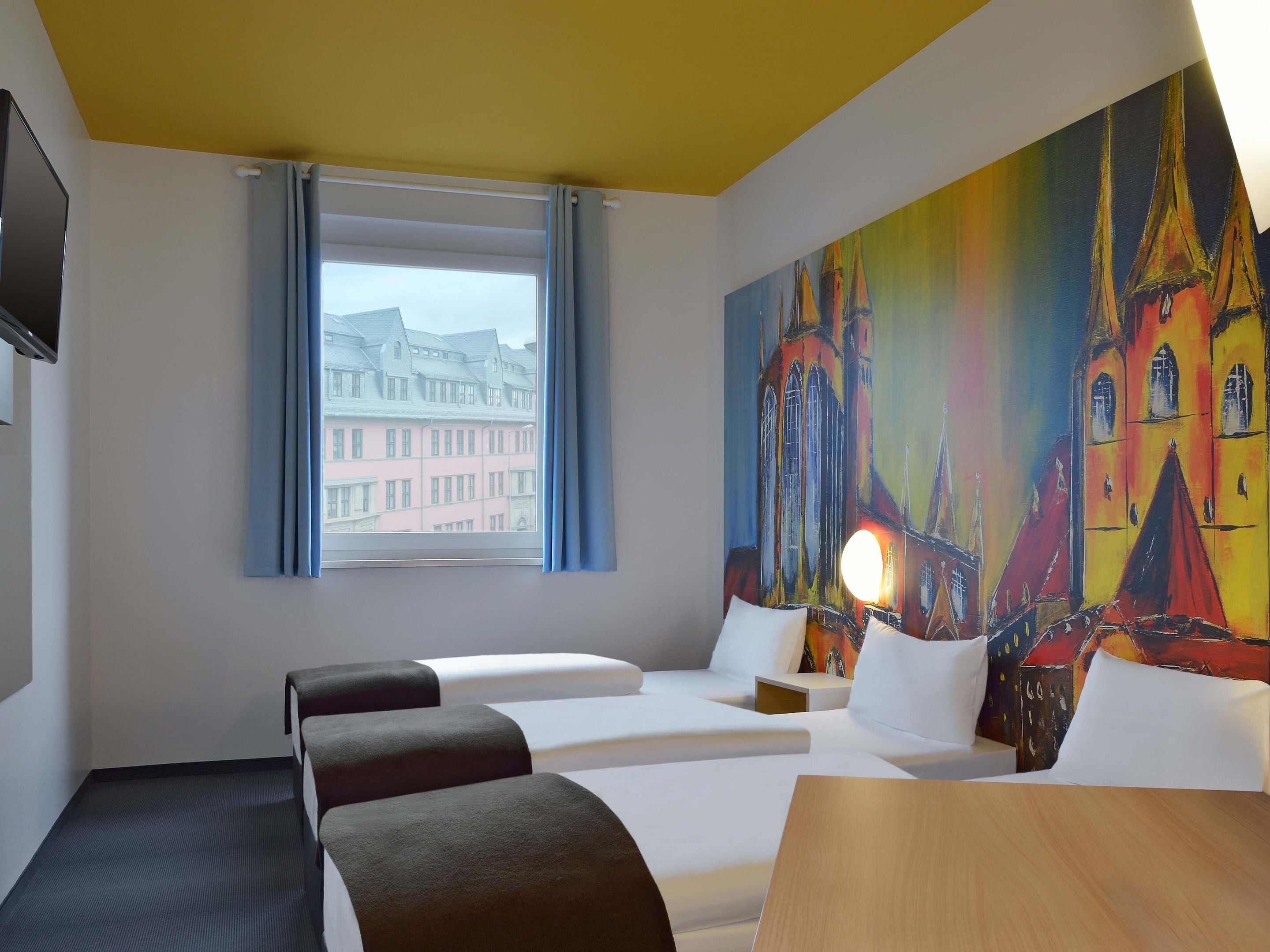 Habitación de hotel con dos camas, una mesa de madera, un cuadro de un edificio colorido, una ventana con vistas a edificios y un televisor de pantalla plana en la pared.