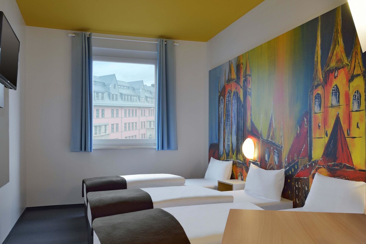 A hotel room with two beds, a wooden table, a painting of a colorful building, a window with a view of buildings, and a flat-screen TV on the wall.