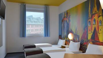 Habitación de hotel con dos camas, una mesa de madera, un cuadro de un edificio colorido, una ventana con vistas a edificios y un televisor de pantalla plana en la pared.