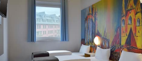 A hotel room with two beds, a wooden table, a painting of a colorful building, a window with a view of buildings, and a flat-screen TV on the wall.